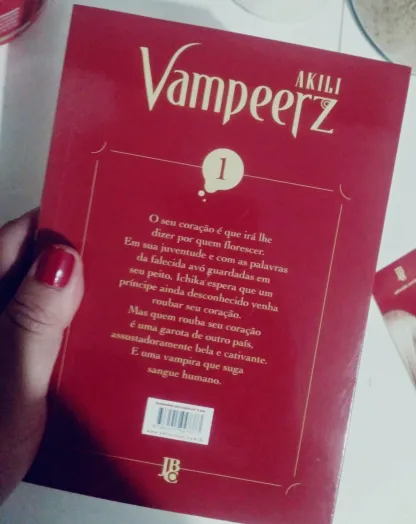 Capa traseira do mangá "Vampeerz" volume 1, com fundo vermelho e texto dourado e branco. Uma mão com unhas pintadas de vermelho segura o livro. Há um resumo da história, destacando o romance entre uma jovem e uma vampira.