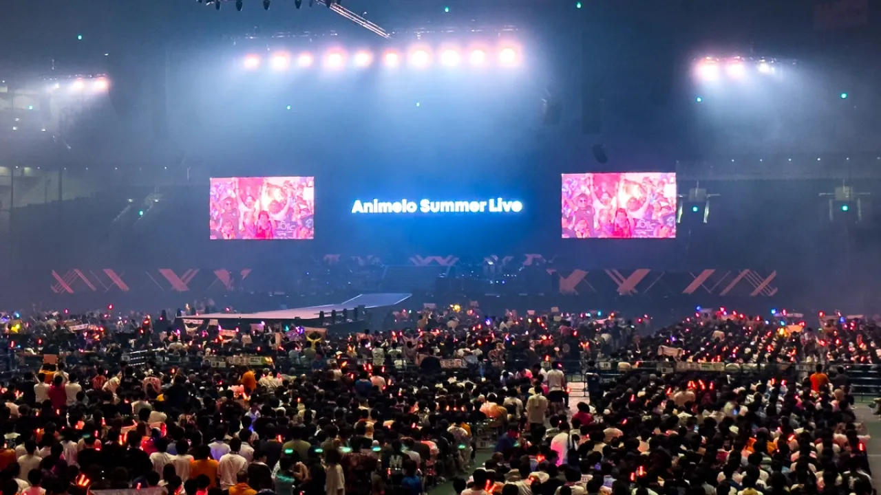 Imagem de um grande show indoor com palco iluminado e três telões. O telão central exibe “Animelo Summer Live”. A plateia está cheia e os fãs seguram bastões de luz, criando um ambiente vibrante típico de concertos de música anime