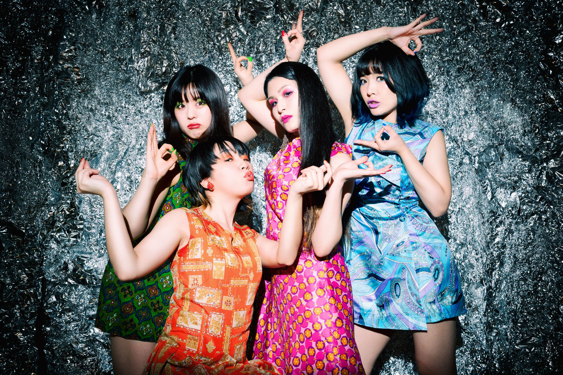 Quatro integrantes da banda Otoboke Beaver posam diante de um fundo metálico texturizado. Cada uma veste roupas coloridas e estampadas, inspiradas em trajes asiáticos tradicionais. As poses são expressivas e teatrais, com destaque para gestos das mãos e maquiagem marcante, criando uma composição visual vibrante e artística.