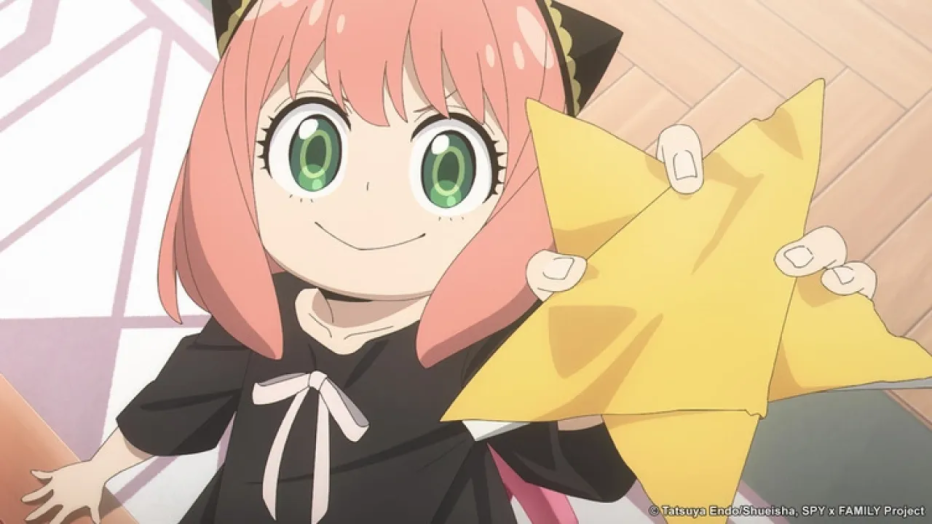 Anya Forger, personagem do anime Spy x Family, aparece sorrindo enquanto segura uma grande estrela amarela. Ela tem cabelo rosa, olhos verdes e veste uma roupa preta com laço branco. O fundo mostra um chão de madeira e uma almofada rosa