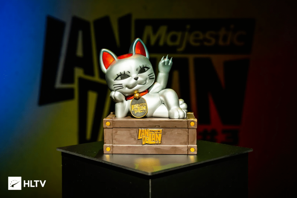 Estatueta de um gato da sorte prateado, com detalhes vermelhos e medalhão dourado no pescoço escrito “Majestic LAN”. O gato está reclinado sobre uma caixa de madeira estilizada com cantos metálicos. Ao fundo, há um painel colorido com o nome “Majestic LAN”, indicando um evento de jogos ou tecnologia.