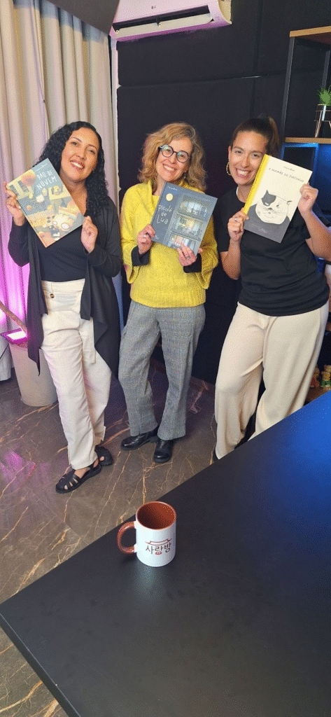 Três pessoas estão em pé, sorrindo e segurando livros. Estão em um ambiente interno com parede escura, cortina à esquerda e chão de mármore. Na frente, há uma mesa com uma caneca branca com interior vermelho e a palavra "Atenea".