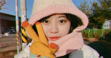 Pessoa ao ar livre usando gorro rosa claro, jaqueta branca e luva amarela com desenho de personagem. Faz gesto de paz com a mão. Céu azul, árvores e construções ao fundo. Atmosfera alegre e ensolarada.